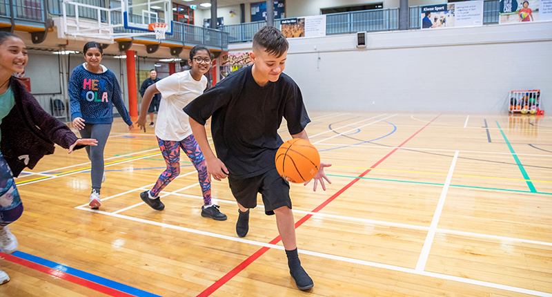 A team of young people chase a player dribbling a basketball inside the stadium at the Allan Brewster Leisure Centre.